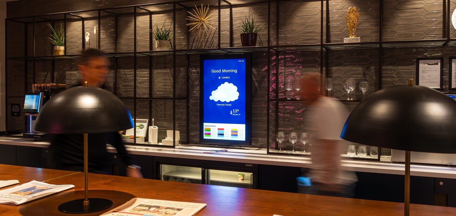 Two residents passing the coffee bar in a first-floor resident lounge, where a portrait Abode screen shows the morning weather and London tube status.