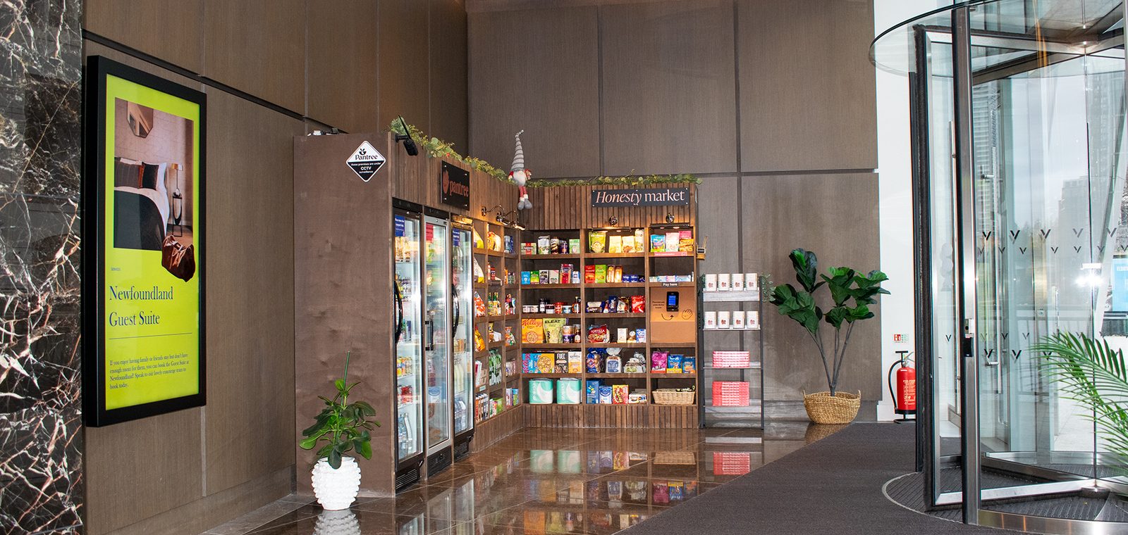 A stocked resident pantry in a residential lobby: chilled cabinets and shelves of packaged food and drink, with a portrait Abode screen on the wall a few steps away.
