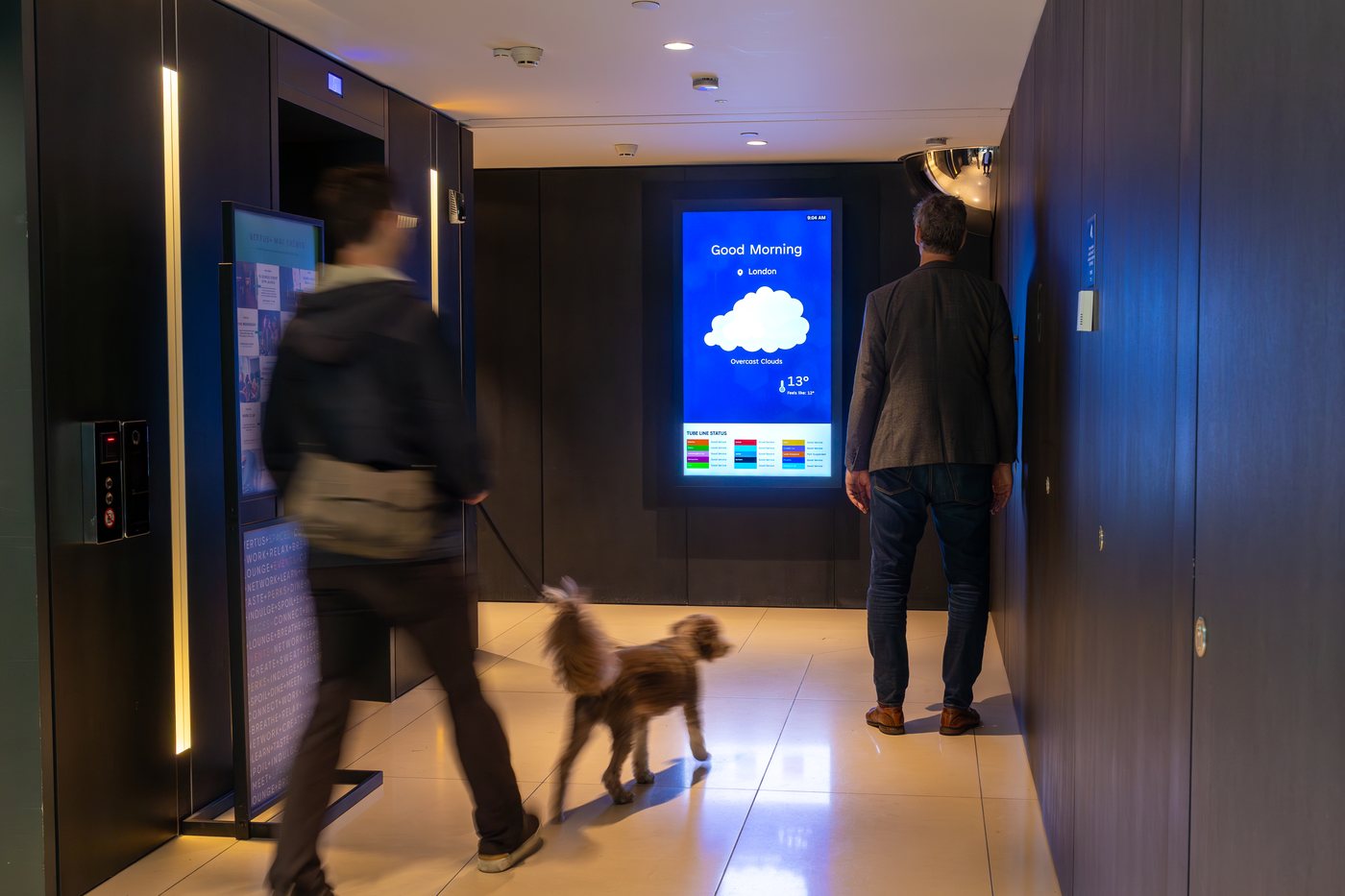 A resident walking a dog through a dark timber-lined Canary Wharf lift lobby, past an Abode screen showing London weather and tube status.