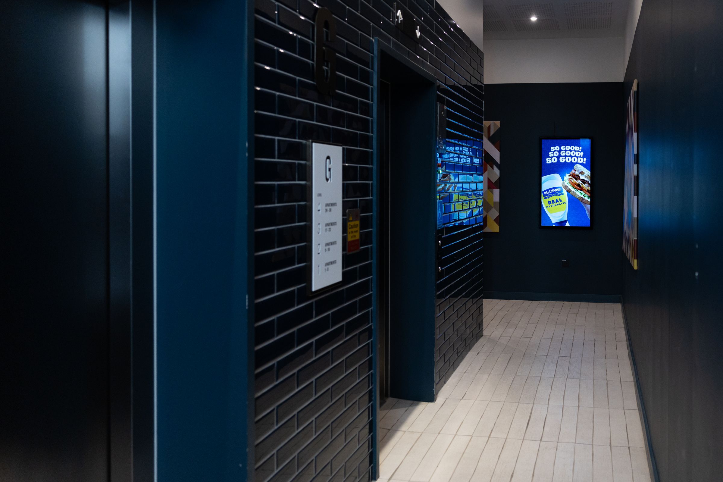 A navy glazed-brick residential lift lobby with portrait Abode screens glowing down the corridor.
