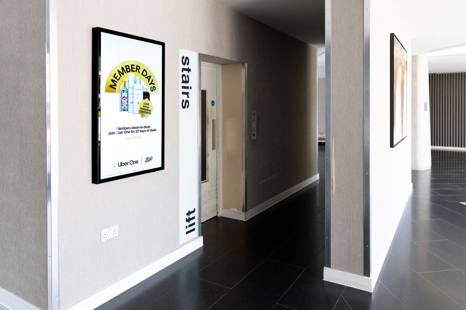 A bright residential corridor with stairs wayfinding on the wall and a portrait Abode screen running a retail offer.
