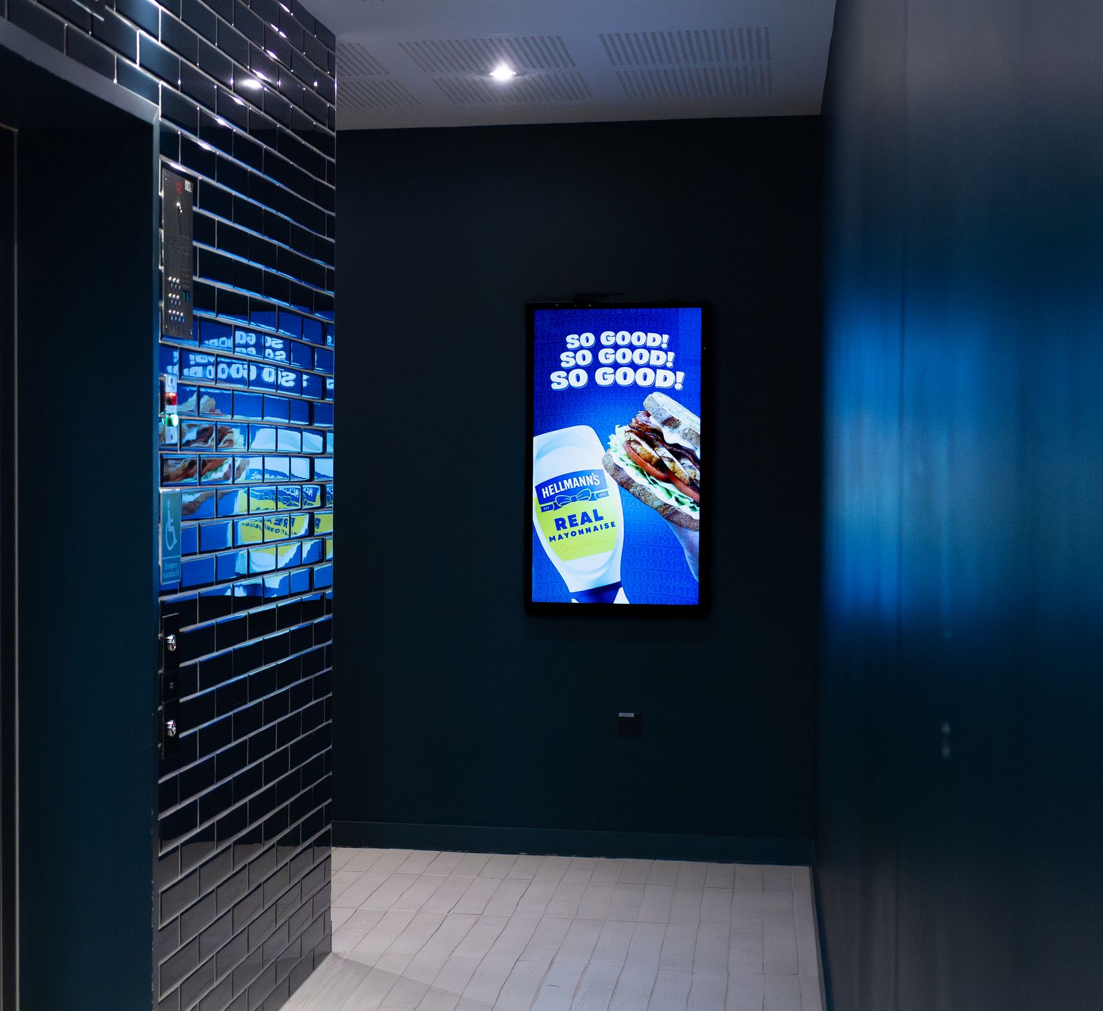 Close view of an Abode lobby screen mounted on navy glazed brick, running a food-brand advert.