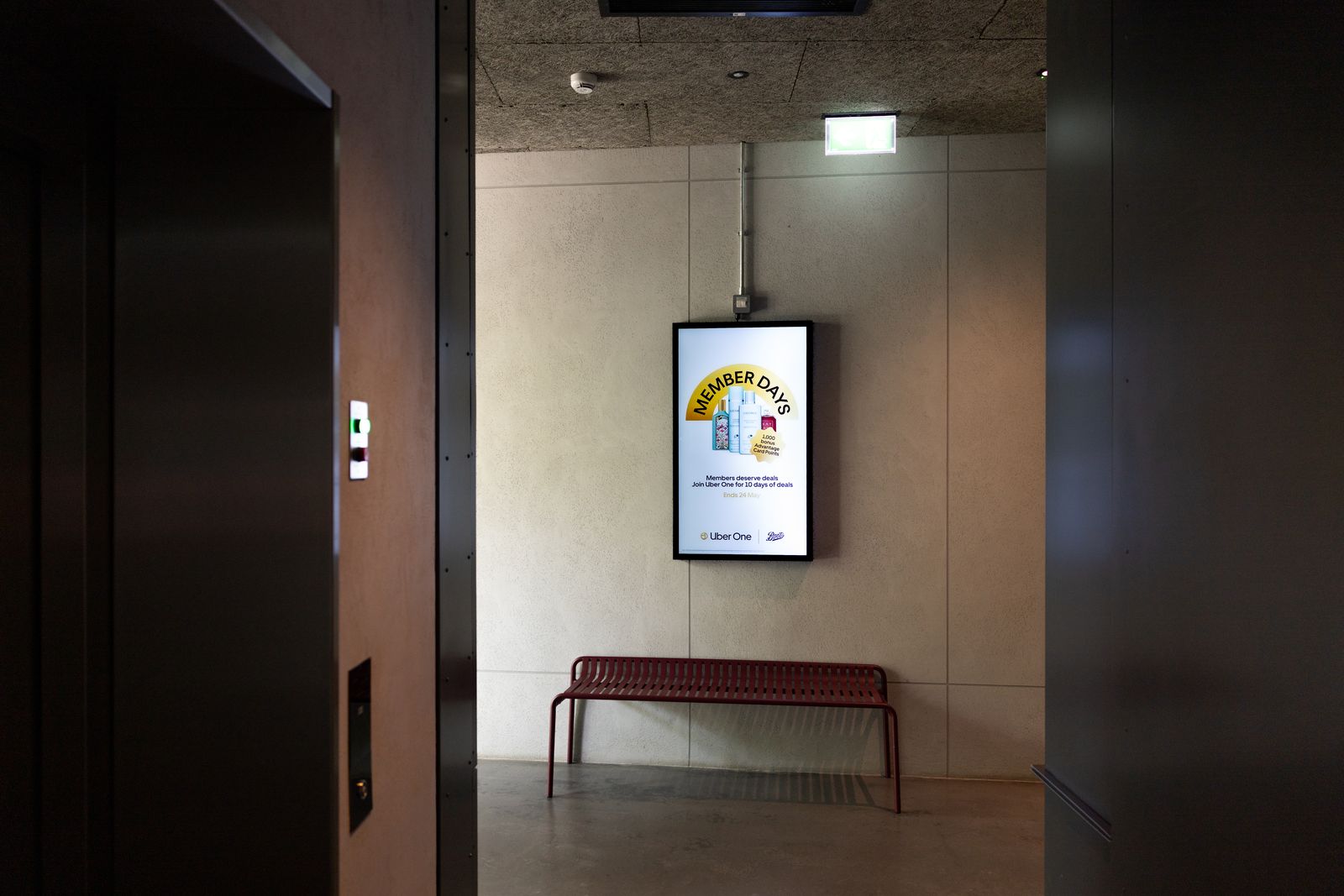 A lift lobby with a concrete ceiling and a red bench; a portrait Abode screen glows beside the lift doors.